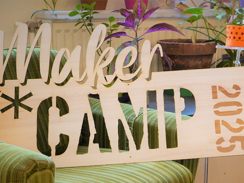 Holzschild mit ausgeschnittenem Schriftzug „Maker * Camp 2025“, auf einem grünen Sessel platziert, mit Pflanzen und Bastelmaterialien im Hintergrund.: 