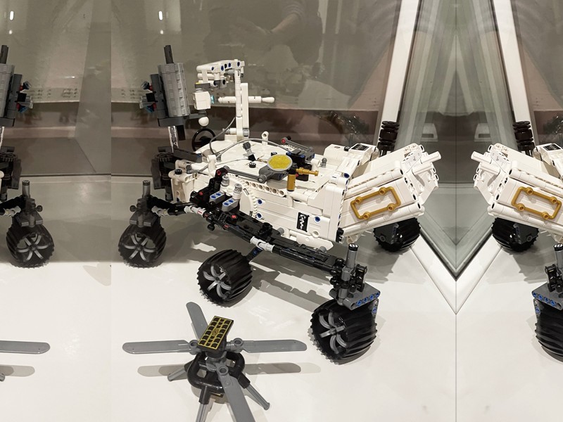 Ein LEGO-Modell eines Mars-Rovers und einer kleinen Drohne, ausgestellt in einer Museumsvitrine, mit einer Spiegelung auf einer Glasoberfläche.: 