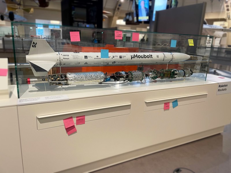 A model rocket displayed in a glass case with several colorful sticky notes attached to the case and base, located in a museum exhibit labeled "Raketen" (Rockets).: Feedback on and Suggestions for the Rocket Display