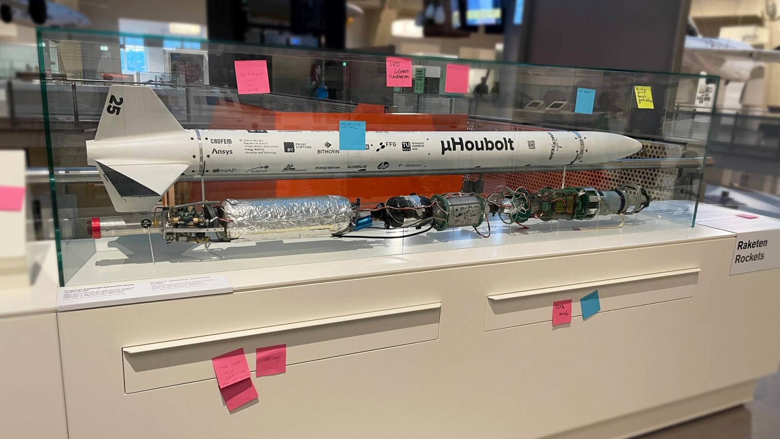 A model rocket displayed in a glass case with several colorful sticky notes attached to the case and base, located in a museum exhibit labeled "Raketen" (Rockets).: Feedback on and Suggestions for the Rocket Display