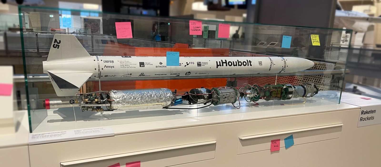 A model rocket displayed in a glass case with several colorful sticky notes attached to the case and base, located in a museum exhibit labeled "Raketen" (Rockets).: Feedback on and Suggestions for the Rocket Display