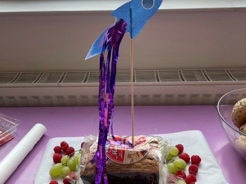 A loaf cake decorated with a paper rocket and surrounded by grapes and raspberries, placed on a purple table near a window.: Cake-rocket? Rocket-cake? Either way, the reunion after summer was celebrated with cake and rockets!