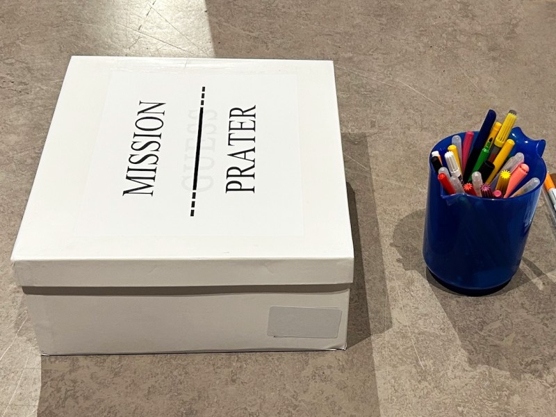 Eine weiße Box mit der Aufschrift "MISSION PRATER" auf dem Boden, daneben ein blauer Becher mit bunten Markern und Stiften sowie weiteres Schreibmaterial.: In der „MISSION PRATER“-Box werden Ideen und Anliegen der Jugendlichen gesammelt.