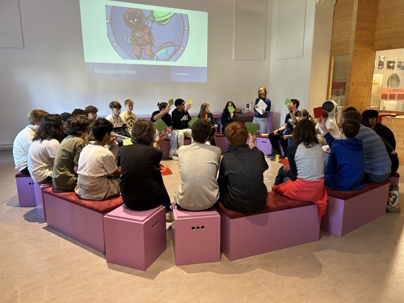 A group of people sitting in a circle on purple benches, attending a presentation in a museum, with an image projected on the wall.: Voting and assessment of “fun facts” and facts.