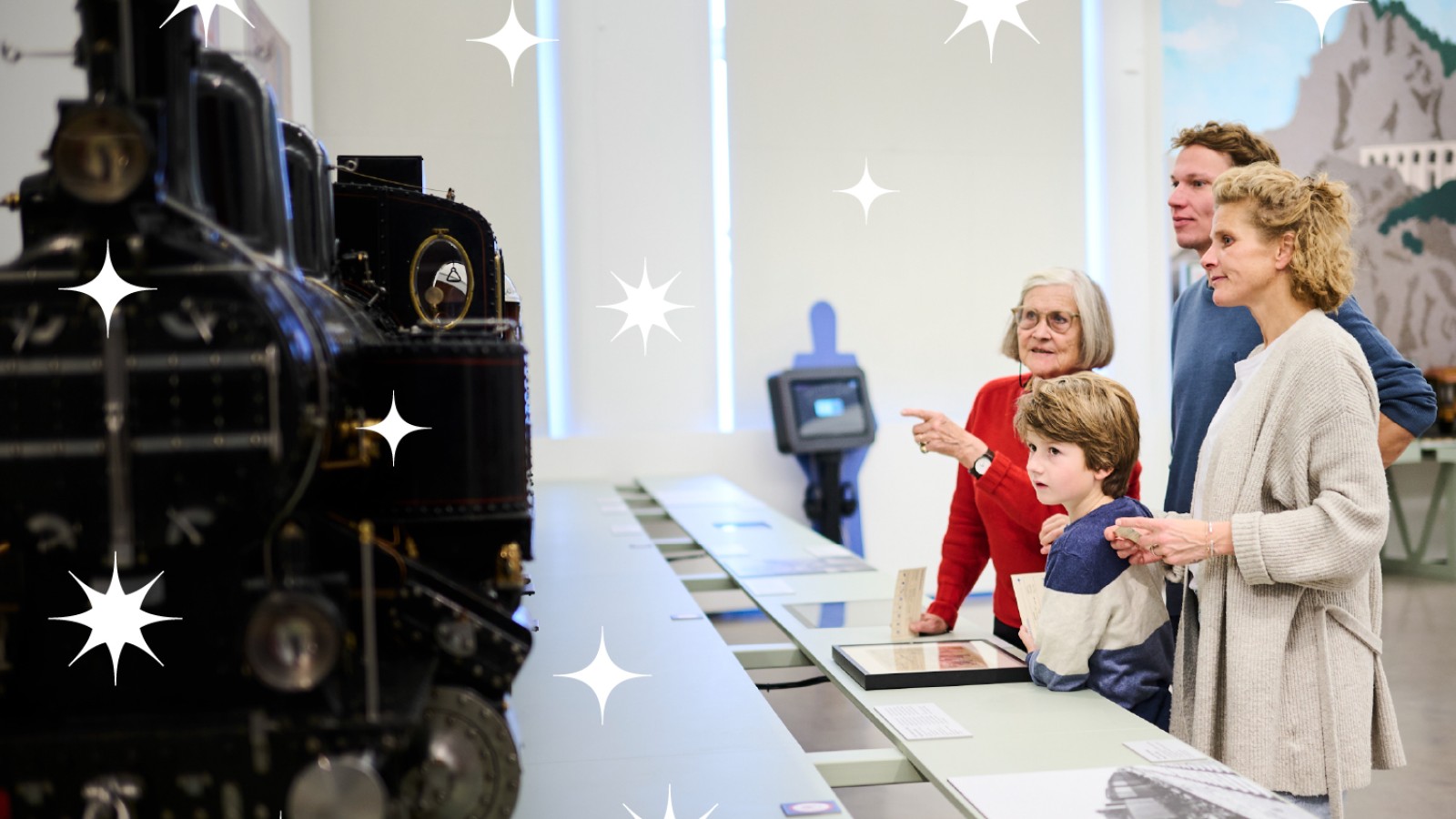 A group of four people — two adults, an older adult, and a child — stands in an exhibition room in front of a large black locomotive model, looking at the exhibits; decorative white star shapes are scattered across the image.: