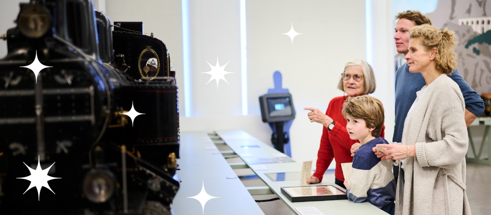 A group of four people — two adults, an older adult, and a child — stands in an exhibition room in front of a large black locomotive model, looking at the exhibits; decorative white star shapes are scattered across the image.: