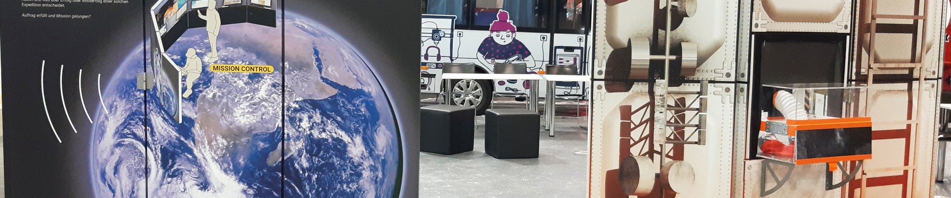 Zwei große, interaktive Ausstellungskulissen in einem Weltraumthema: eine mit "Mission Control" und einem Bild der Erde, die andere zeigt eine futuristische Struktur mit mechanischen Details. Im Hintergrund ist ein Bus mit der Aufschrift "roadLAB" zu sehen.: Aufbau des Settings am Tag vorher …