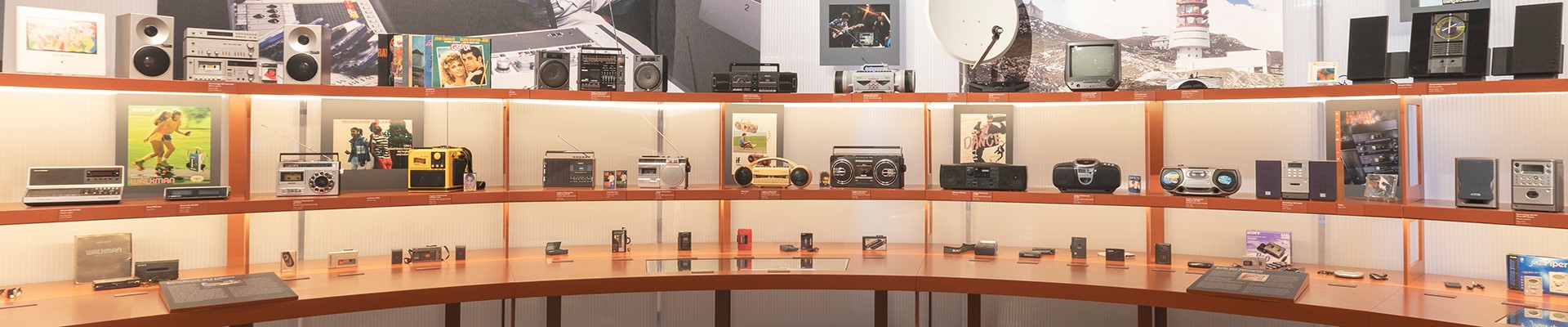 Eine Museumspräsentation im Technischen Museum Wien mit einer Vielzahl tragbarer Audiogeräte, darunter Kassettenrekorder, Boomboxes und Walkmans, auf Regalen angeordnet.: 