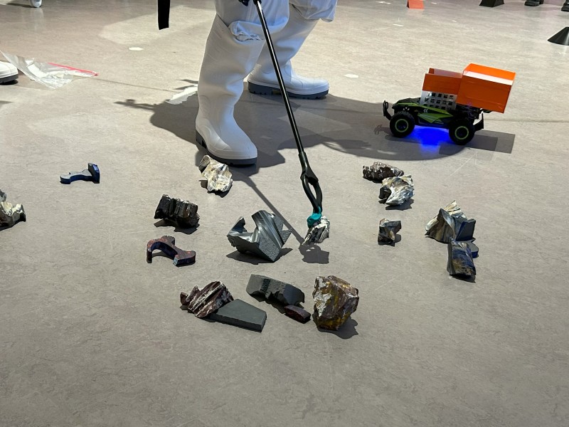 A person in white boots uses a grabber tool to collect simulated rocks on the floor, with a small rover carrying an orange container nearby.: “Mars rover in action: Rock samples are being collected and transported to the habitat.“