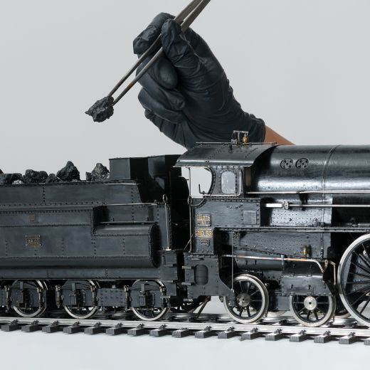 Hand mit schwarzem Handschuh hält eine Pinzette mit Kohle über ein detailreiches Modell einer Dampflokomotive auf Gleisen Im Bann der Bahn