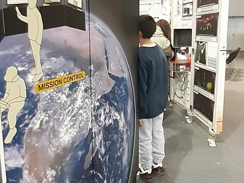 Ein Kind steht neben einem "Mission Control"-Panel mit einem Bild der Erde, Teil einer weltraumbezogenen Ausstellung mit Kontrollbildschirmen und futuristischen Details im Hintergrund.: Hier ist es nur ein kleiner Schritt von der Erde auf den Mars. Die EXPEDITION MARS lädt Besucher_innen zu einem Kurzbesuch ein.