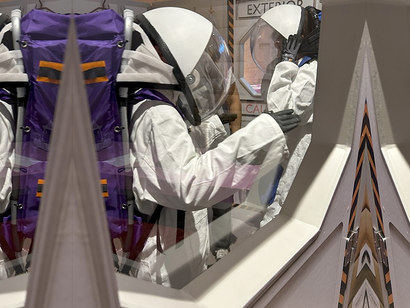  Two individuals wearing white astronaut suits and helmets are viewed through a window, standing in a simulated airlock labeled "EXTERIOR" and "CAUTION.": 