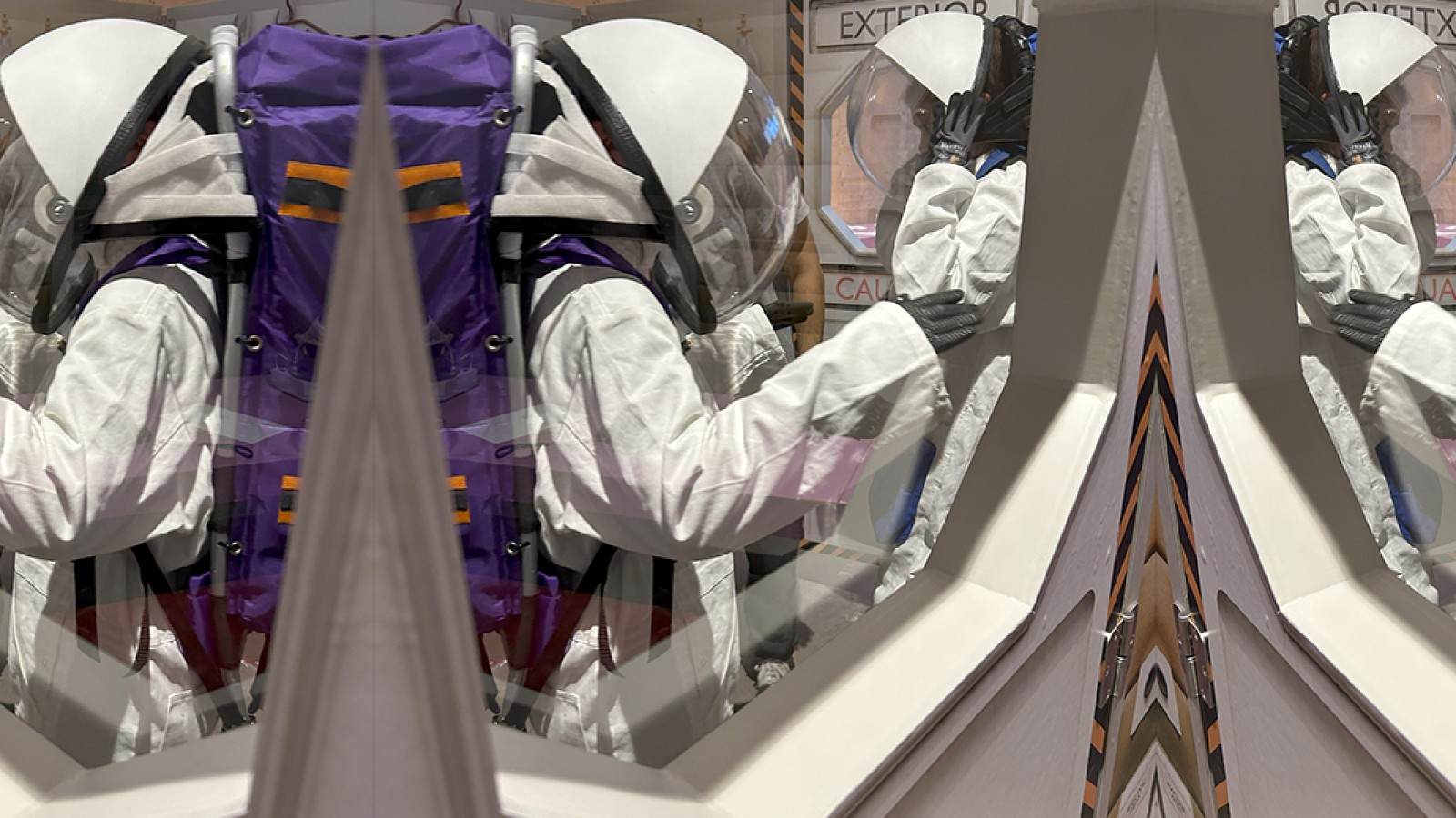  Two individuals wearing white astronaut suits and helmets are viewed through a window, standing in a simulated airlock labeled "EXTERIOR" and "CAUTION.": 