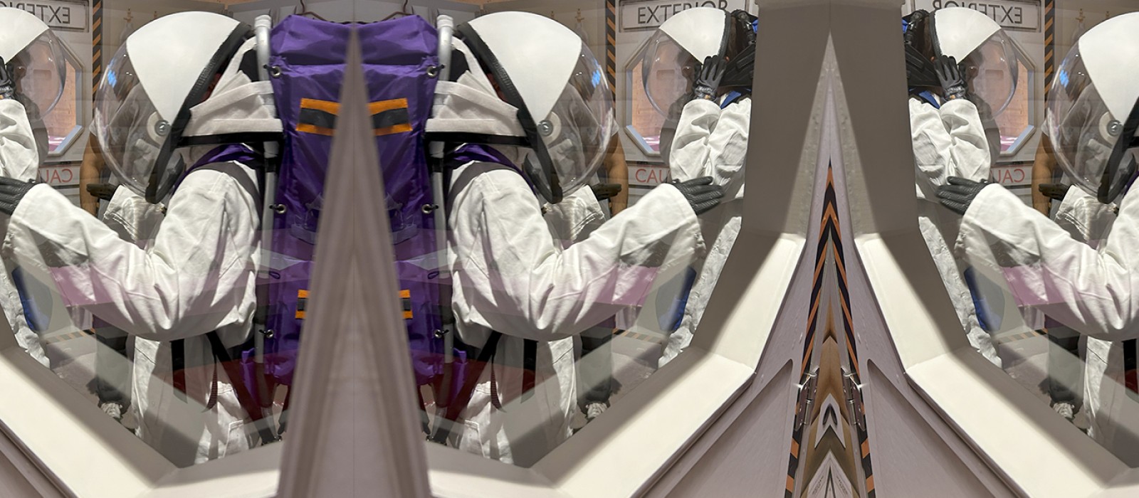  Two individuals wearing white astronaut suits and helmets are viewed through a window, standing in a simulated airlock labeled "EXTERIOR" and "CAUTION.": 