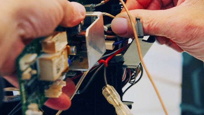 Nahaufnahme der Hände einer Person, die konzentriert an der offenliegenden Elektronik und Verkabelung eines Geräts arbeitet. Man sieht filigrane Drähte, Stecker und Bauteile, während die Finger präzise Handgriffe ausführen. Reparieren statt Wegwerfen? Oft unnötig schwer