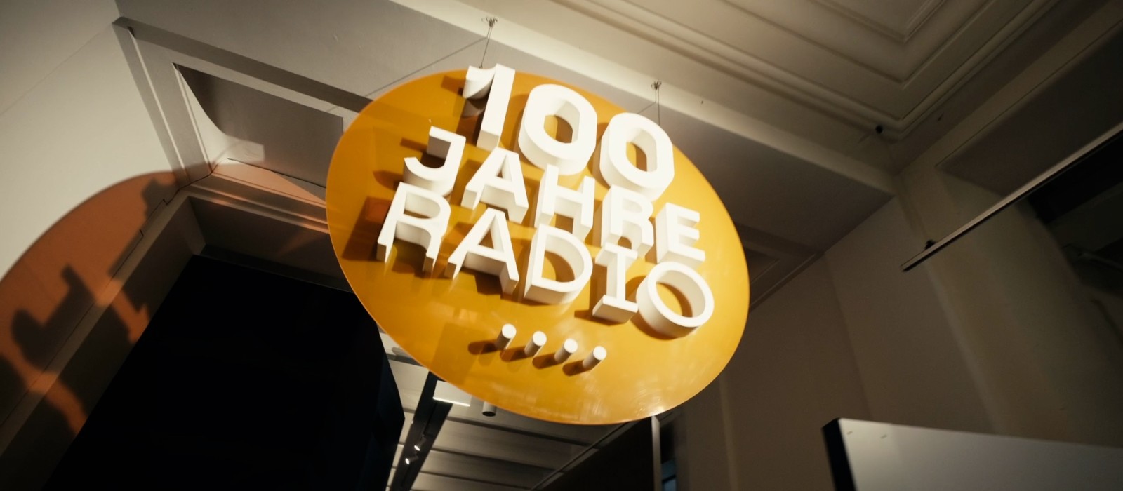 A close-up of a large orange circular sign with "100 Years of Radio" written in bold white letters, hanging from the ceiling.: 