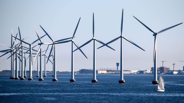 Eine perspektivische Aufnahme eines Offshore-Windparks im tiefblauen Meer. Eine lange Reihe weißer Windkraftanlagen erstreckt sich in einer Kurve von links nach rechts im Bild. Im Vordergrund rechts segelt ein kleines weißes Segelboot an den massiven Turbinen vorbei, während im Hintergrund eine Küstenlinie mit Industriegebäuden und Baukränen unter einem klaren, hellblauen Himmel zu sehen ist. Was macht die Nachhaltigkeit im Museum?