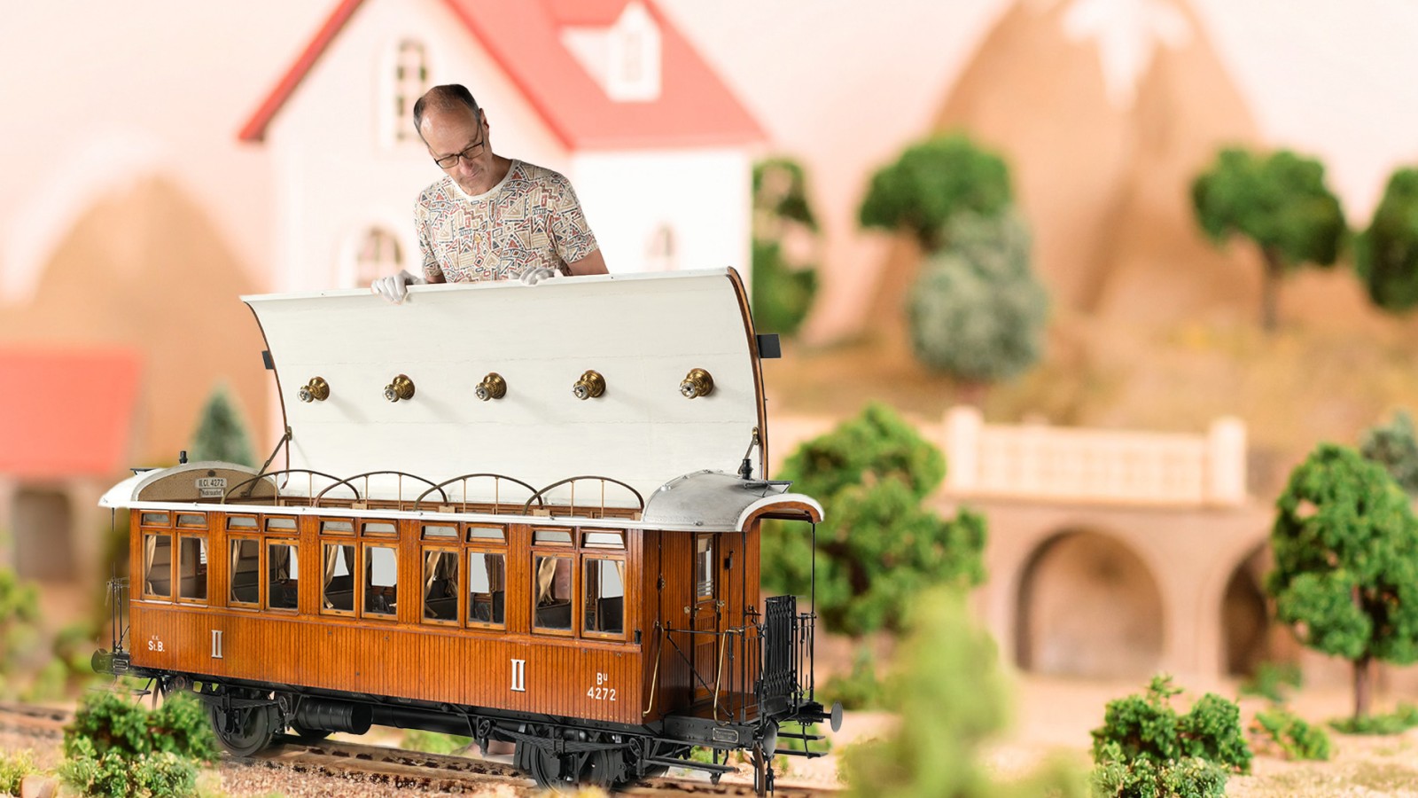 Ein erwachsener Mann mit weißen Handschuhen hebt das Dach eines braunen Modell-Personenwagens an; im Hintergrund sind modellhafte Häuser, Bäume und Berge zu sehen.:
