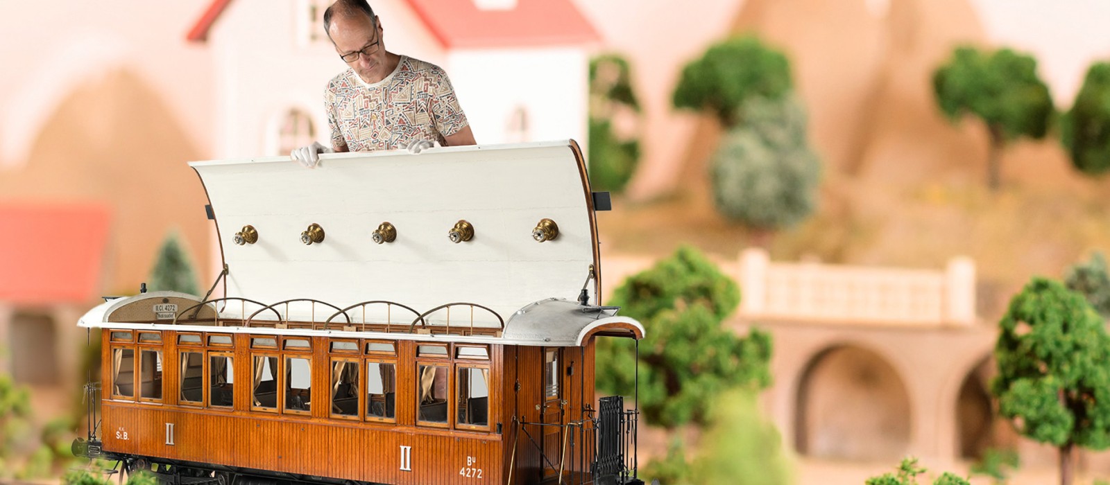 Ein erwachsener Mann mit weißen Handschuhen hebt das Dach eines braunen Modell-Personenwagens an; im Hintergrund sind modellhafte Häuser, Bäume und Berge zu sehen.: