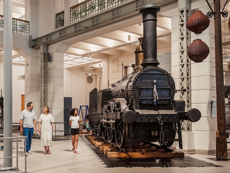 Three people walk through a bright exhibition hall with old machines and a steam locomotive.